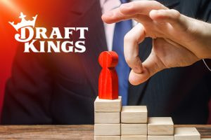 A businessman in a suit flicks a red pawn figure off a stack of wooden blocks, symbolizing a fall from power, with the DraftKings logo prominently displayed in the background.