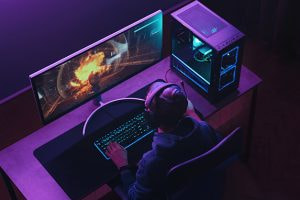 A gamer wearing a headset sits at a desk in a dimly lit room, focused on a large curved monitor displaying an intense in-game explosion scene. The gaming setup includes a high-performance desktop PC with visible cooling fans illuminated by blue and purple lighting.