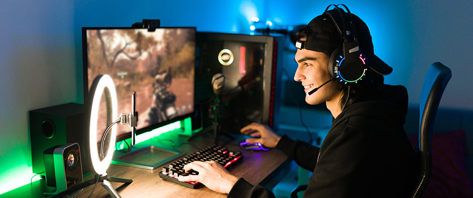 A gamer wearing a headset and a backward cap sits at a desk in a brightly lit room, smiling and focused on a game displayed on a monitor. The setup includes a high-performance desktop PC with colorful lighting, a ring light, and various peripherals illuminated with vibrant colors.