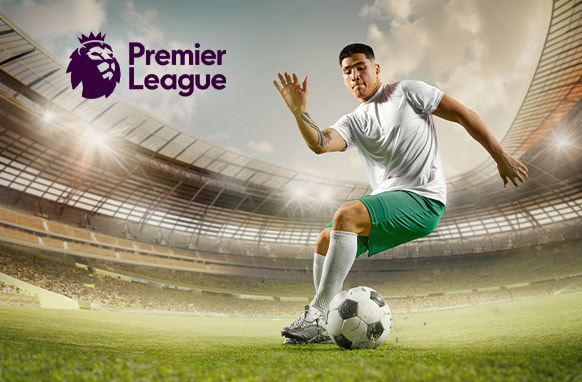 A football player wearing a white shirt and green shorts is in action on the pitch, skillfully controlling the ball inside a large stadium. The Premier League logo is displayed in the upper left corner, with a bright sky and stadium lights in the background, suggesting a Premier League match setting.