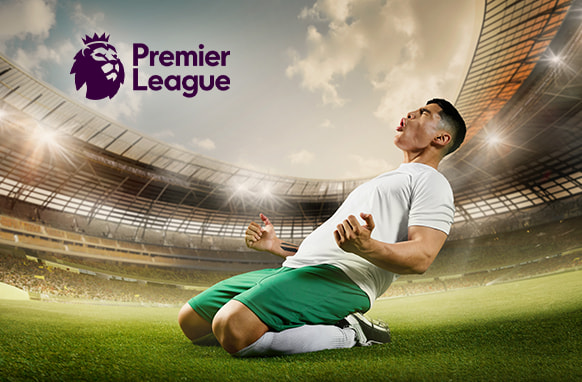 A football player in green shorts and a white shirt celebrates by sliding on his knees in a large stadium under a bright, partly cloudy sky. The Premier League logo is displayed in the top left corner, evoking the excitement of a Premier League match.