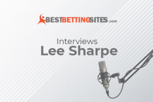 The image shows the logo of "BestBettingSites.com" at the top, followed by the text "Interviews Lee Sharpe" in the center. To the right, there's a professional microphone set up, suggesting that the image is promoting an interview, likely with former footballer Lee Sharpe. The overall design is clean and minimalist, featuring light grey diagonal lines in the background.