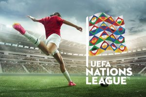 A football player in action on the field, kicking a ball in front of a stadium, with the UEFA Nations League flag displayed prominently in the foreground.