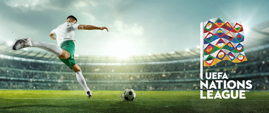 A football player preparing to kick a ball on the field in front of a large stadium, with the UEFA Nations League flag displayed prominently on the right side.