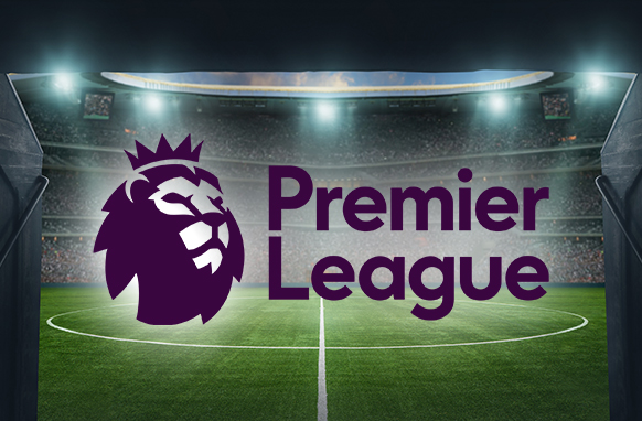 The image shows the Premier League logo featuring a lion's head on a purple background, placed in the centre of a large football stadium. The stadium is well-lit, with empty seats surrounding the football pitch, creating an atmospheric backdrop for a Premier League matchday.