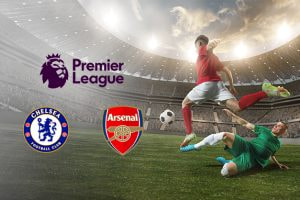 Premier League match between Chelsea and Arsenal with team logos, showing a dramatic scene of a player in red taking a shot on goal as a defender in green slides in to block, set against a packed stadium background.