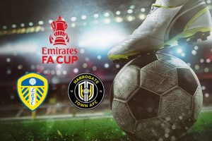 A close-up of a football on a pitch under stadium lights, with a player's foot poised on top of the ball. The image prominently features the Emirates FA Cup logo, along with the Leeds United and Harrogate Town AFC crests, symbolising their upcoming match in the tournament.